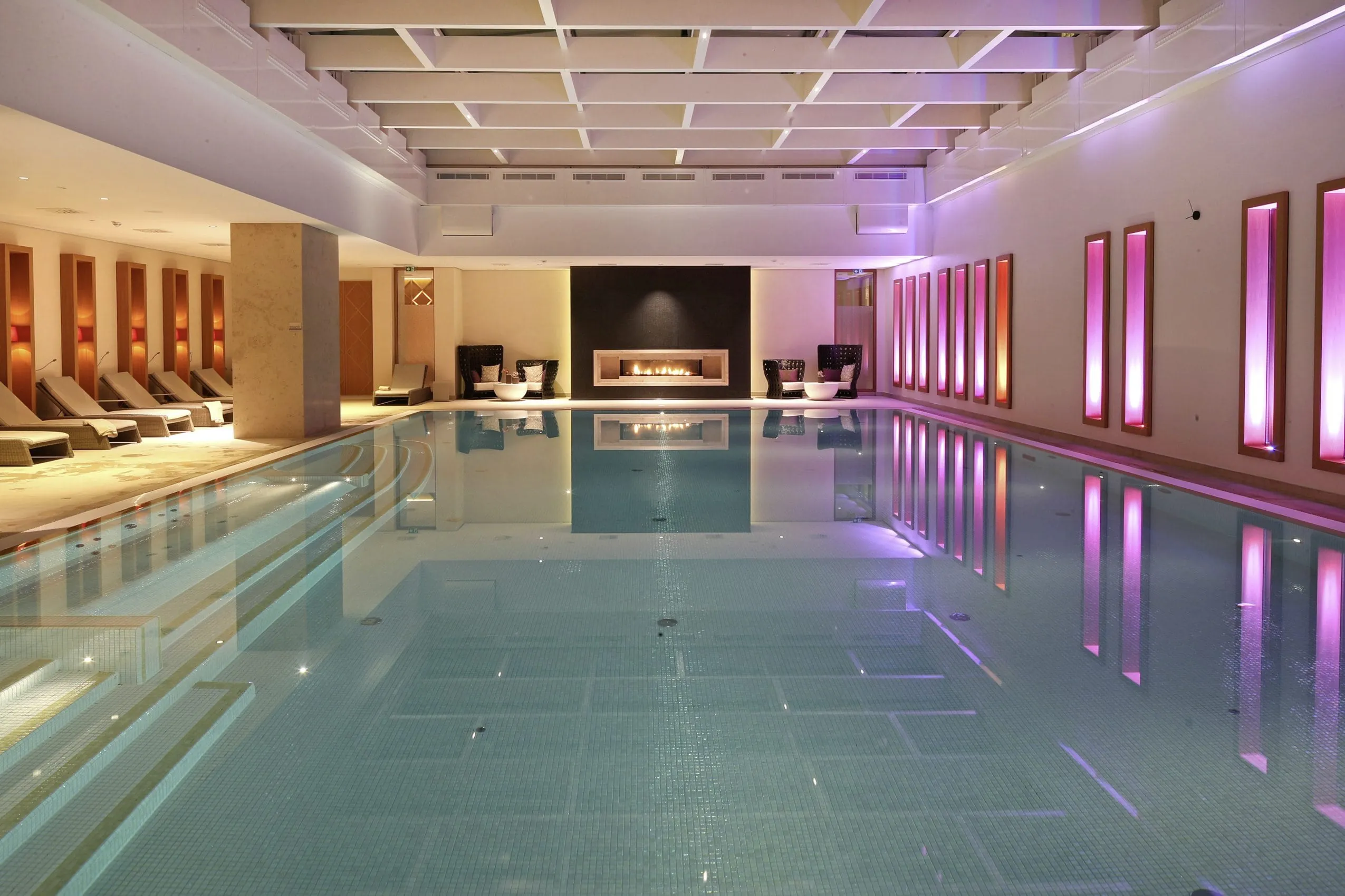 Gro&szlig;er Indoorpool mit strahlend t&uuml;rkis-blauem Wasser. Leuchten links und rechts vom Pool sorgen f&uuml;r stimmungsvolles Licht. Um den Pool herum sind zahlreiche Ruheliegen mit weichen Polstern.