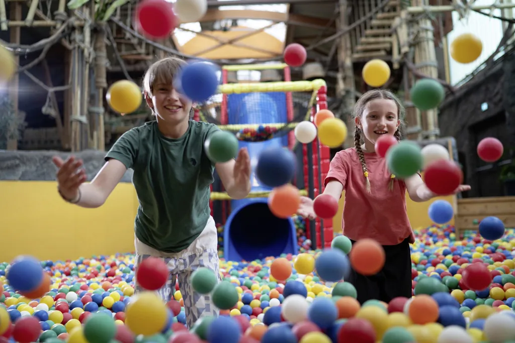 Eine Junge und ein Mädchen stehen im Bällebad und werfen bunte Bälle in Richtung Kamera. Im Hintergrund ist der Klettergarten und eine blaue Röhrenrutsche zu sehen.