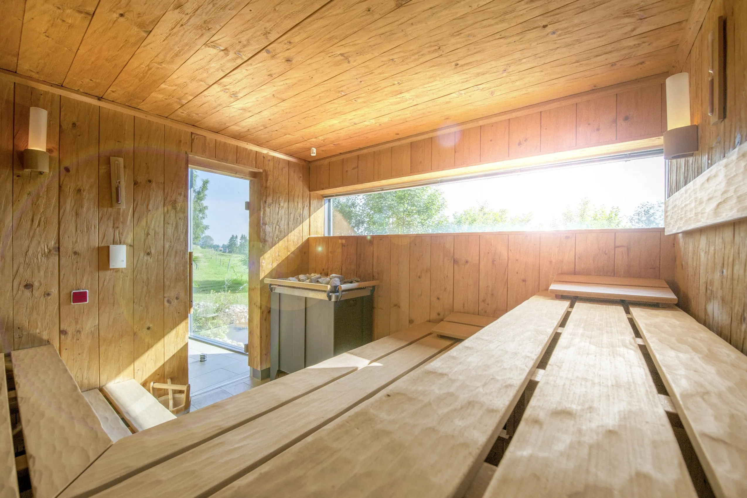 Sauna aus hellem Holz mit schmalem, langen Fenster durch das die Sonne scheint.