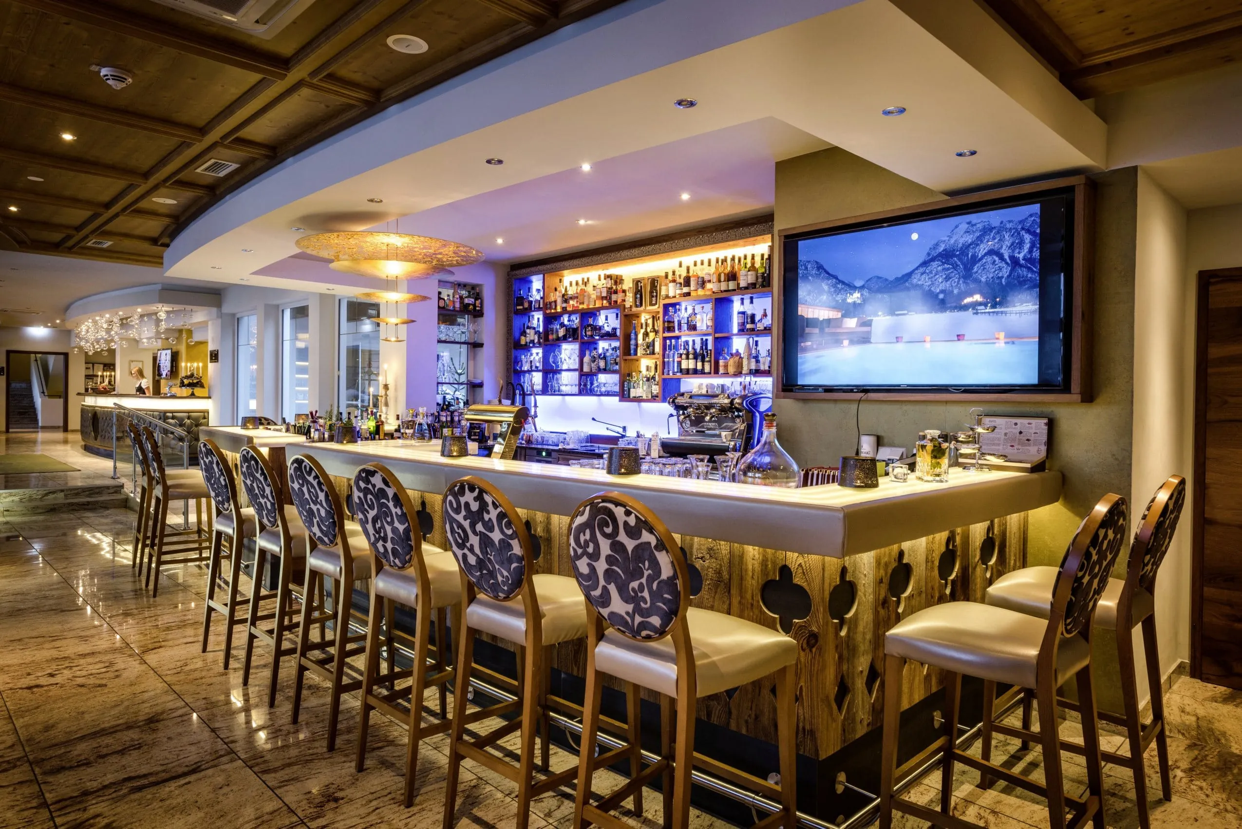 Bar mit rustikaler Holzverkleidung und blau-wei&szlig; gemusterten Barhockern. Die Spirituosen im Wandregal der Bar sind durch blaue Beleuchtung stilvoll in Szene gesetzt.