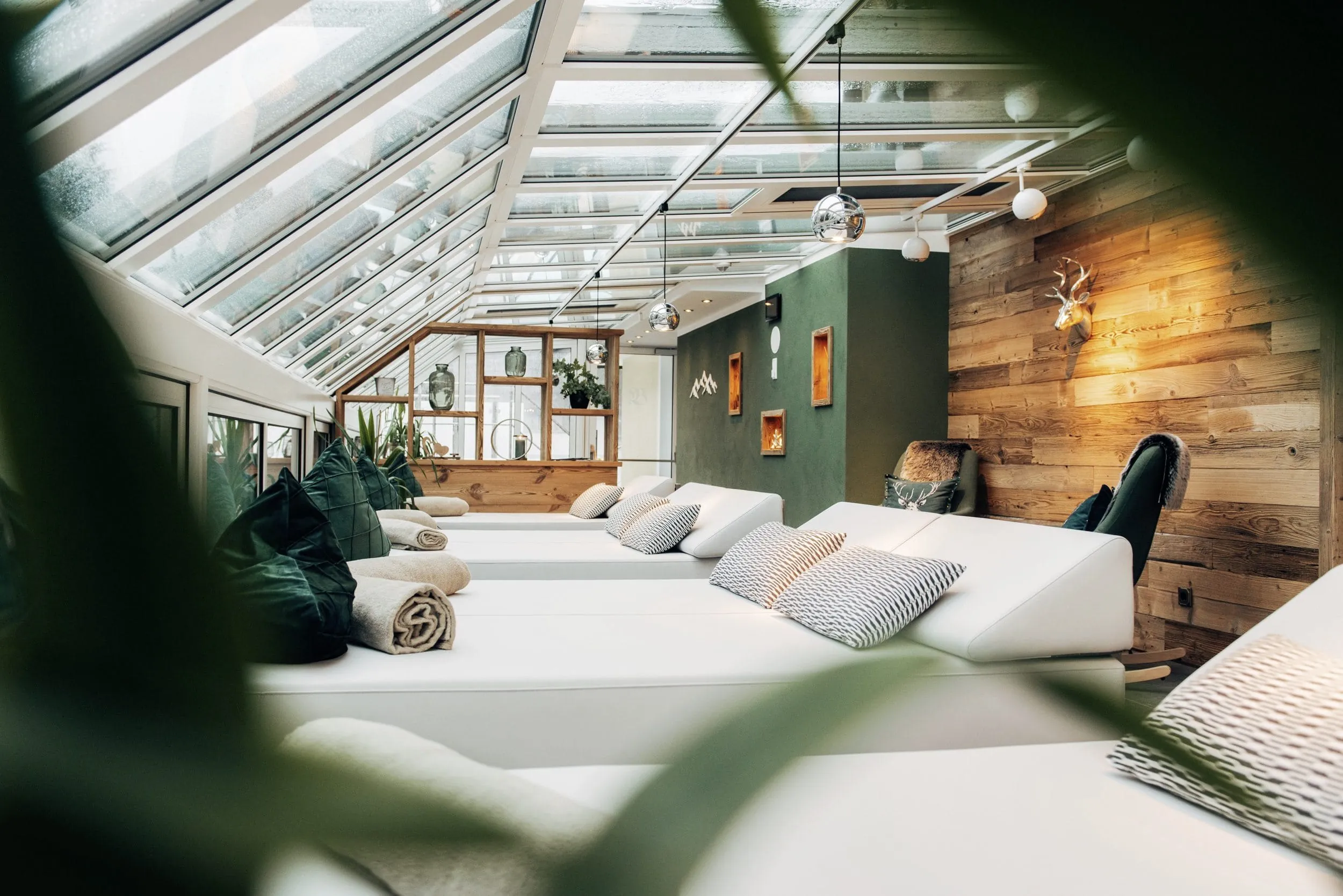 Zahlreiche Daybeds in hellem grau nebeneinander mit Kissen und Decken f&uuml;r Gem&uuml;tlichkeit in einem verglasten Wintergarten mit Blick nach drau&szlig;en in die Natur.
