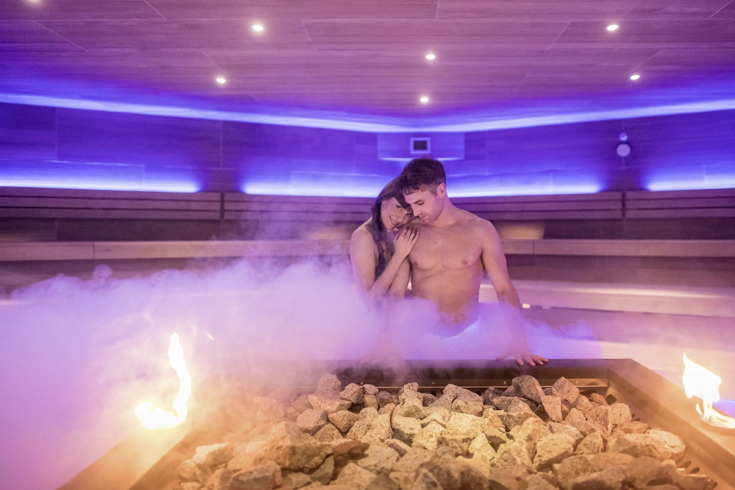 Pärchen kuschelt in der großzügigen Sauna mit stimmungsvoller Beleuchtung in Blau- und Lilatönen und Dekoflammen.