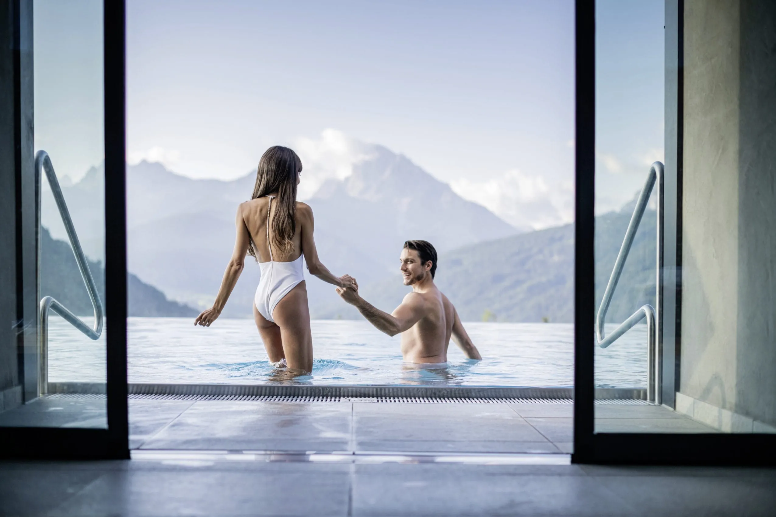 Mann geleitet Frau in weißem Badeanzug die Pooltreppe des Alpen Tesitin hinunter. Im Hintergrund sieht man das Felsmassiv der Dolomiten.