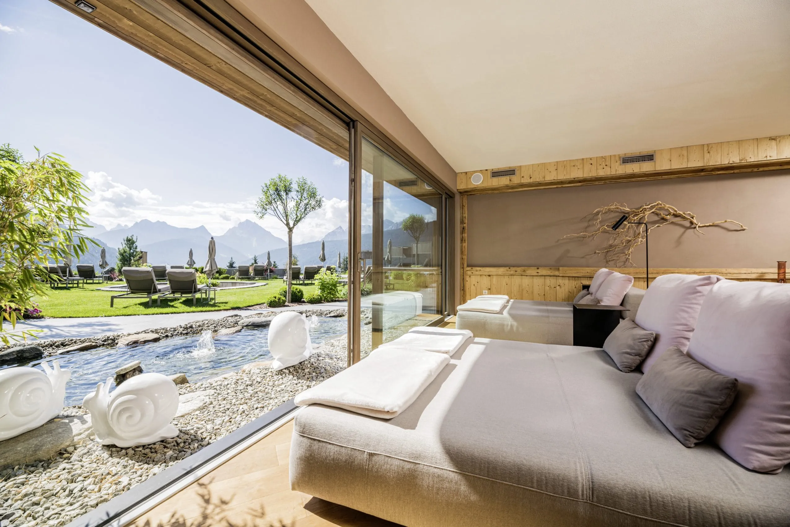 Ruheraum im Alpen Tesitin mit naturfarbenen Ruheliegen und Blick auf den Sprudelteich vor dem Fenster