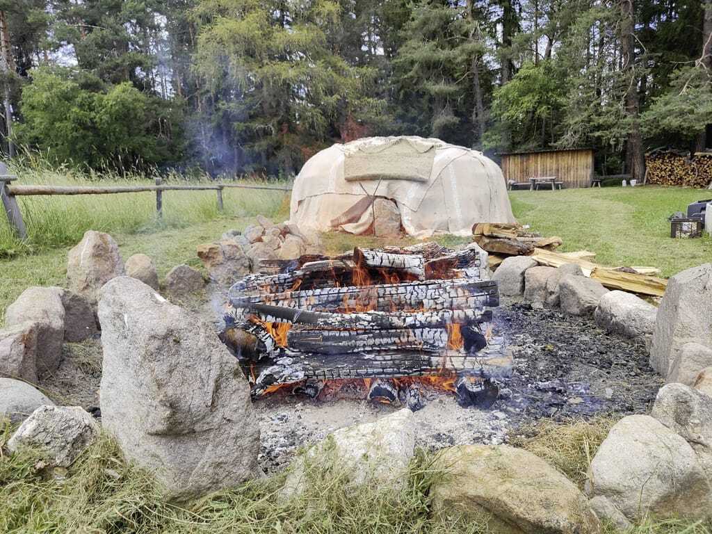Natursauna im Wald des Naturhotel Pfösl mit offenem Feuer und ein mit Decken umhülltes Weidengebäude im Hintergrund