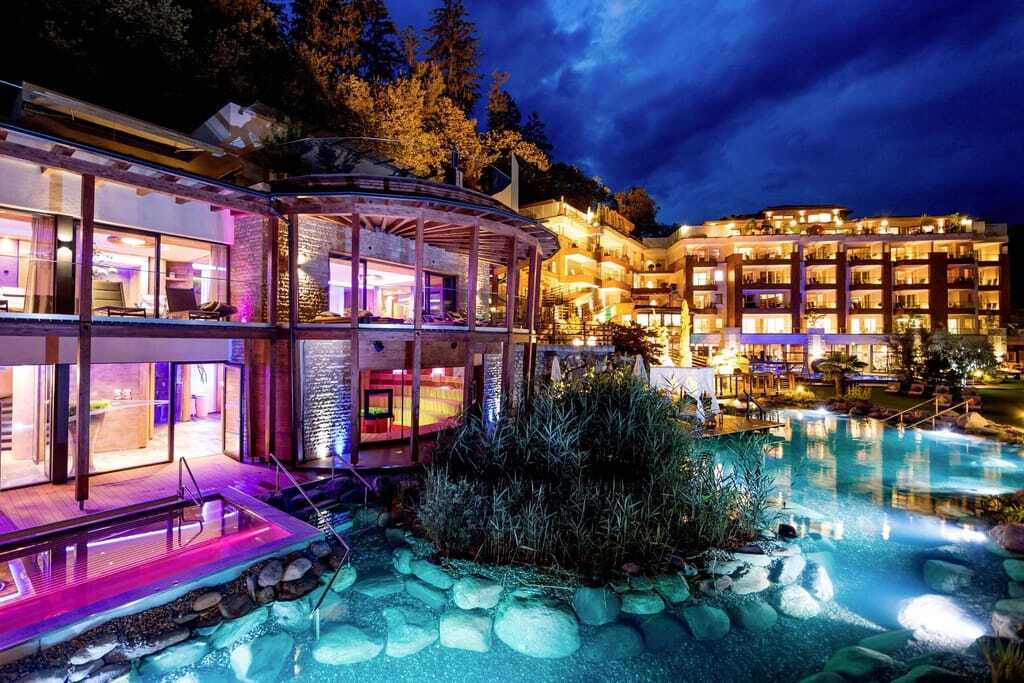 Außenansicht des Quellenhof Luxury Resort Passeier mit beleuchtetem Badeteich, moderner Architektur und Blick in die See-Event-Sauna bei Nacht