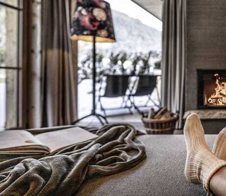 Gemütliches Wohnzimmer der Südtirol Chalets Valsegg mit Kamin, Decke, Buch, Wollsocken und Blick auf verschneite Landschaft