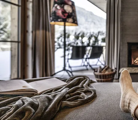 Gemütliches Wohnzimmer der Südtirol Chalets Valsegg mit Kamin, Decke, Buch, Wollsocken und Blick auf verschneite Landschaft