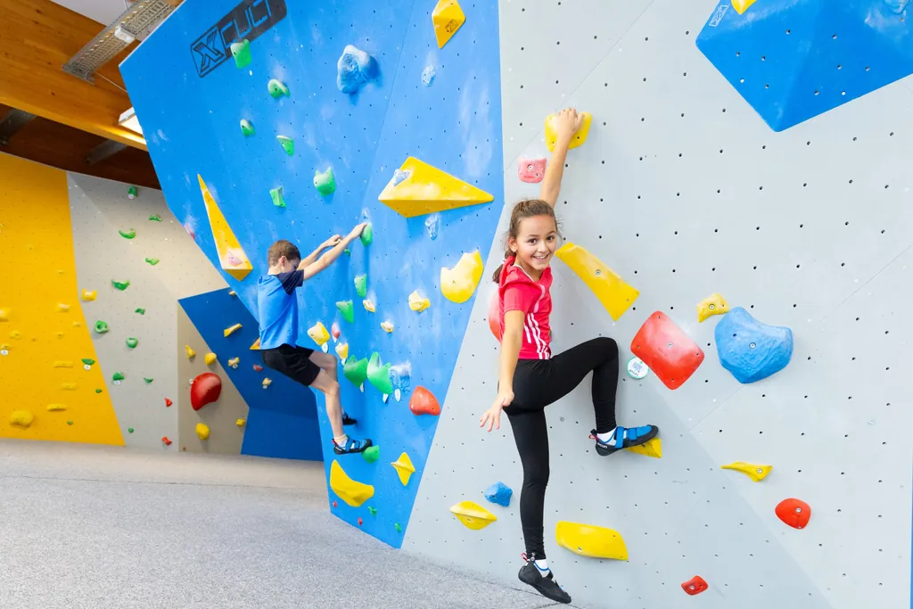 Junges Mädchen und Junge in Sportklamotten an einer Kletterwand. Das Mädchen lächelt in die Kamera, der Junge ist hochkonzentriert am klettern.