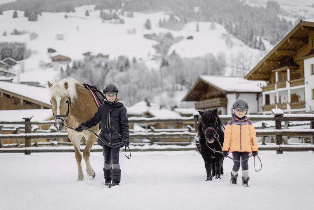 Mädchen neben Pony und Mädchen neben Pferd halten Strick und stehen auf der verschneiten Reitanlage des Bauernhof Hotel Oberschwarzach