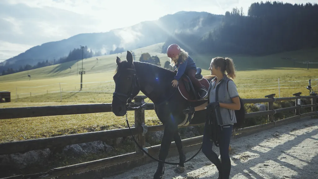 M&auml;dchen auf einem braunen Pferd wird von Reitlehrerin gef&uuml;hrt auf der Au&szlig;enreitanlage des Bauernhof Hotel Oberschwarzach