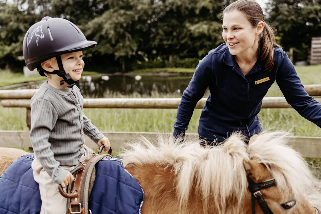 kleiner Junge mit Reithelm sitzt l&auml;chelnd auf einem hellbraunen Pony und Reitlehrerin des Familotel Landhaus Averbeck steht l&auml;chelnd daneben