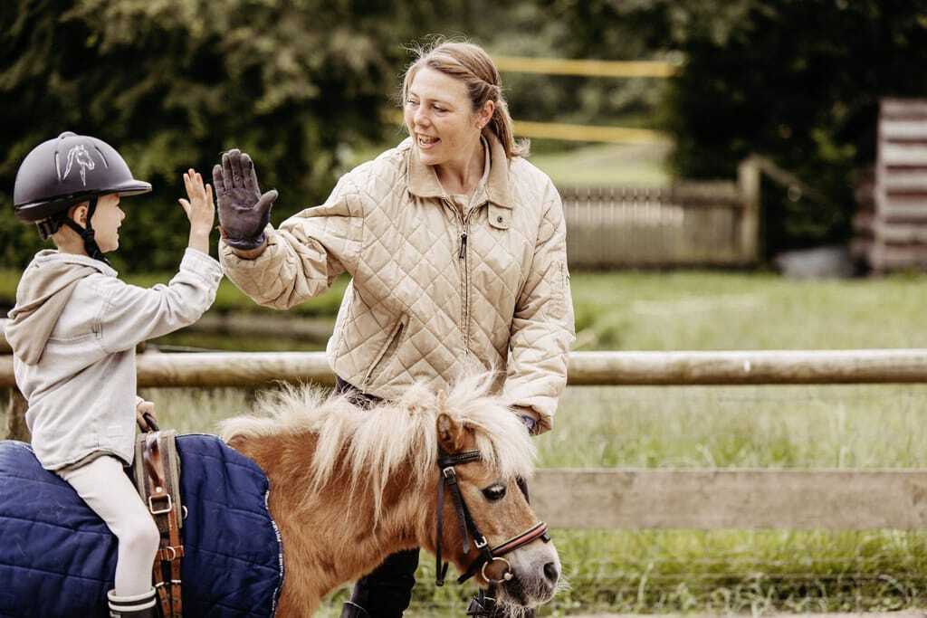 Reitlehrerin gibt kleinem Jungen auf Pony high-five auf dem Außenreitplatz des Familotel Landhaus Averbeck