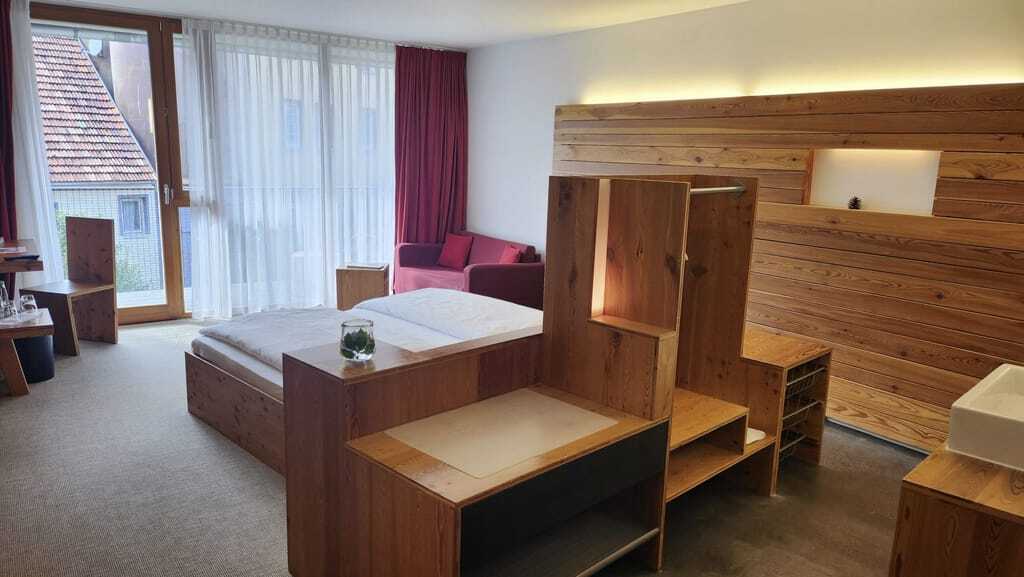 Blick in ein schlichtes Doppelzimmer des Hotel Speidel’s BrauManufaktur mit Holzeinrichtung, Doppelbett und rotem Sofa im Hintergrund