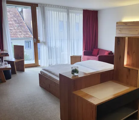 Blick in ein schlichtes Doppelzimmer des Hotel Speidel’s BrauManufaktur mit Holzeinrichtung, Doppelbett und rotem Sofa im Hintergrund