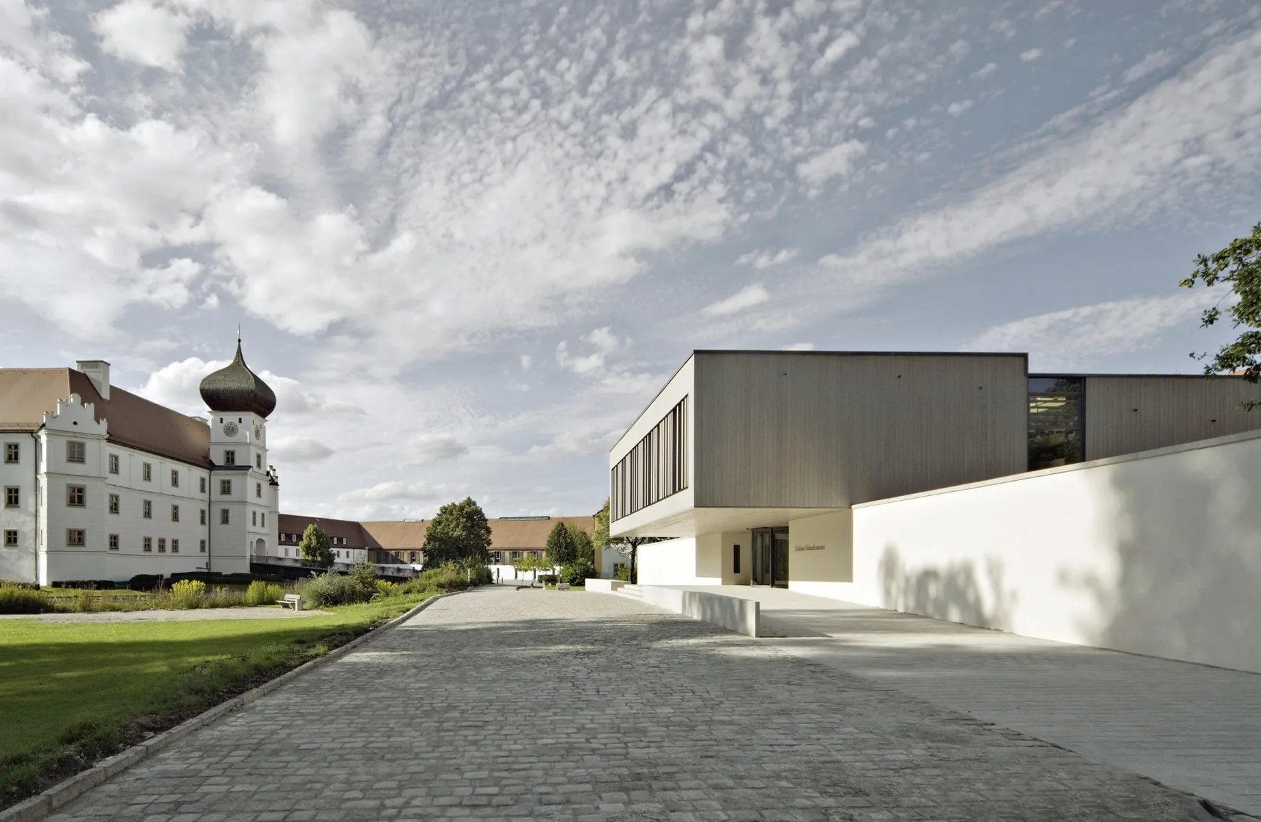 Moderne Fassade des Schloss Hohenkammer und Schlossgebäude mit Turm, dazwischen ein Kopfsteinpflasterweg und blauer Himmel