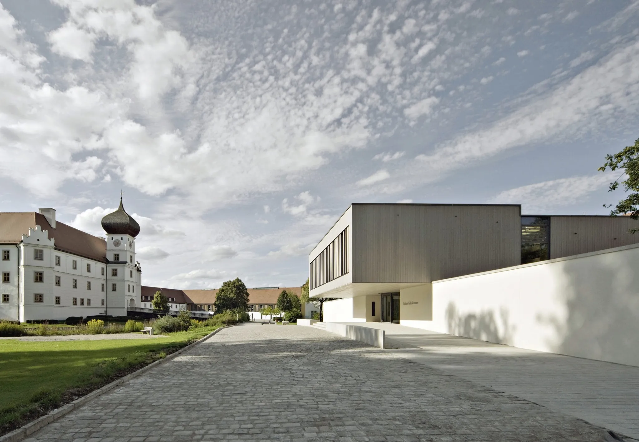 Moderne Fassade des Schloss Hohenkammer und Schlossgebäude mit Turm, dazwischen ein Kopfsteinpflasterweg und blauer Himmel
