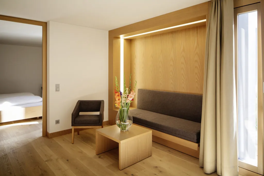 Blick in ein Zimmer des Schloss Hohenkammer mit gepolsterter Sitzbank, Polsterstuhl, kleinem Holz-Beistelltisch mit Blumenvase und Doppelbett im Hintergrund