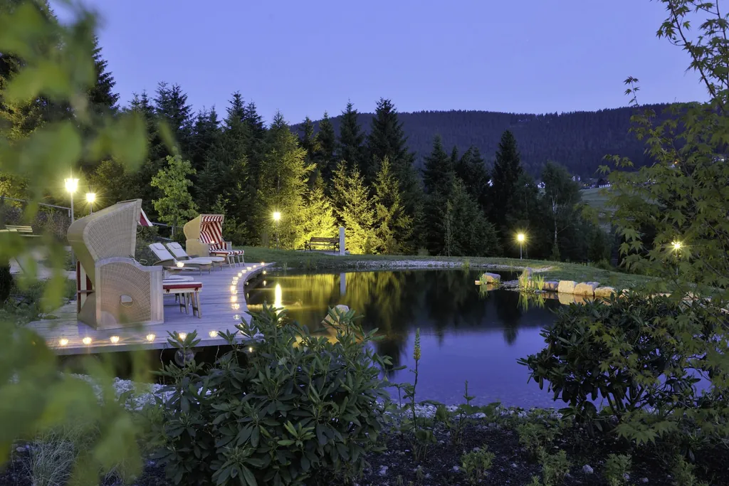 Beleuchteter Naturteich des Hotel Bareiss mit Strandkörben, Liegen und Blick auf das umliegende Schwarzwald-Panorama bei Abenddämmerung
