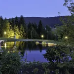 Beleuchteter Naturteich des Hotel Bareiss mit Strandkörben, Liegen und Blick auf das umliegende Schwarzwald-Panorama bei Abenddämmerung