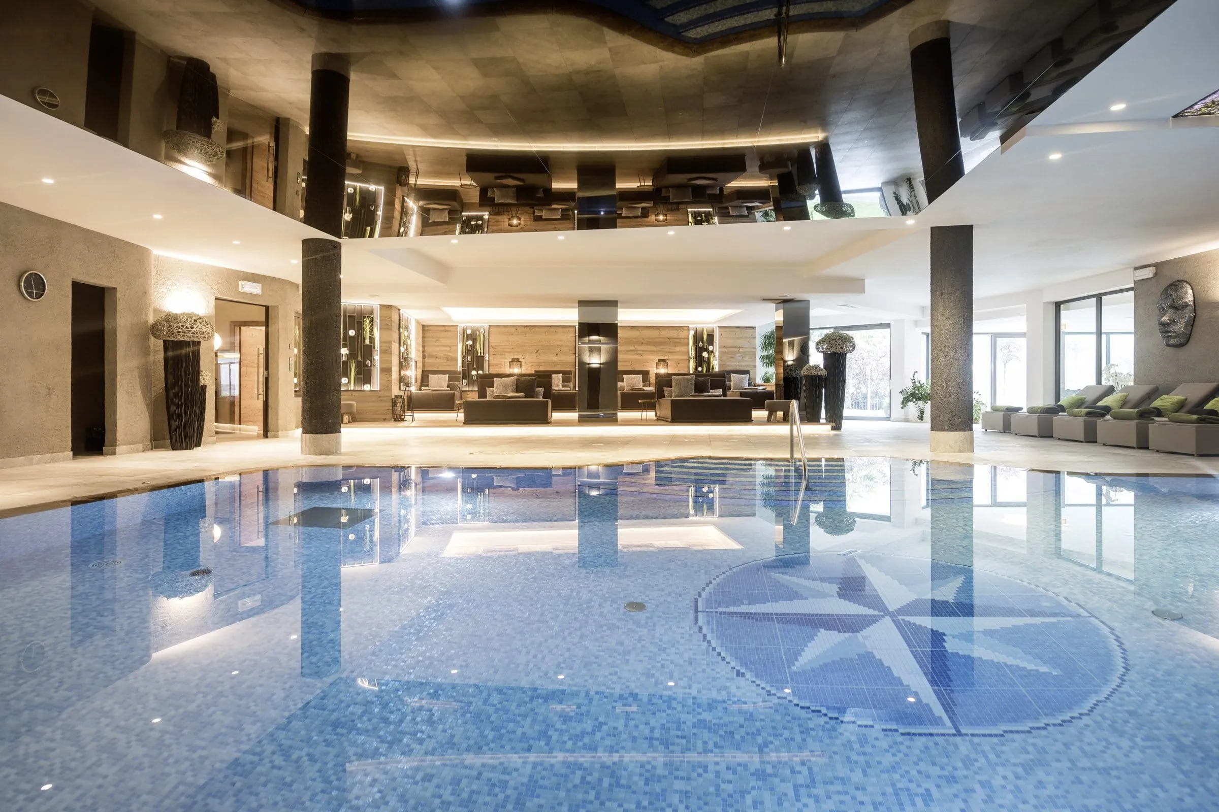 Indoorpool des Wellnesshotels in Südtirol Das Alpenschlössel mit grauen Liegen an der Seite, edlen dunklen Säulen und einer verspiegelten Decke.