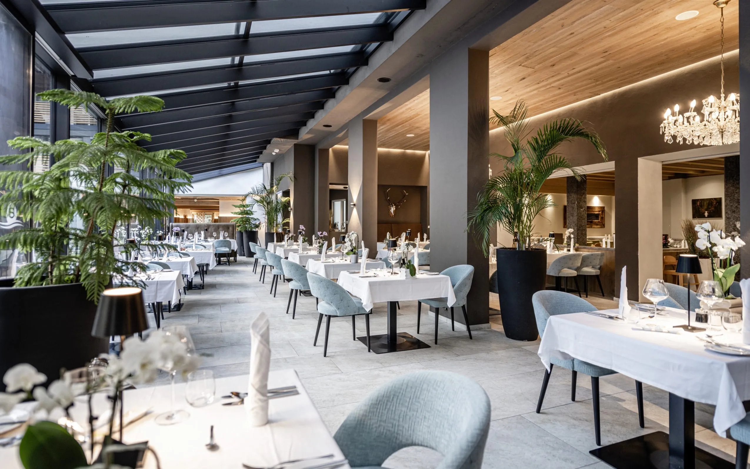 Edel eingerichtetes Restaurant des Stroblhofs mit Wintergarten. Schicke Kronleuchter, weiße Tischdecken, gedeckt-blaue Polsterstühle und Stoffservietten