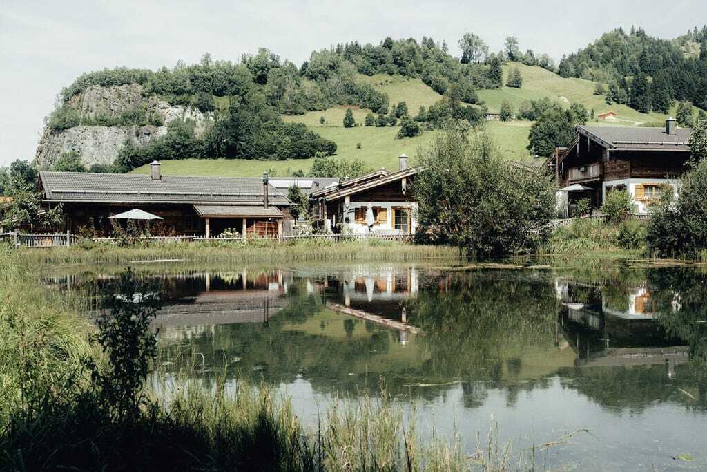 Grüne hügelige Wiesen mit kleinen Wäldchen und Büschen. Im Vordergrund ein kleiner smaragdgrüner See an dem gemütliche, kleine Chalets in rustikaler Holzoptik stehen.