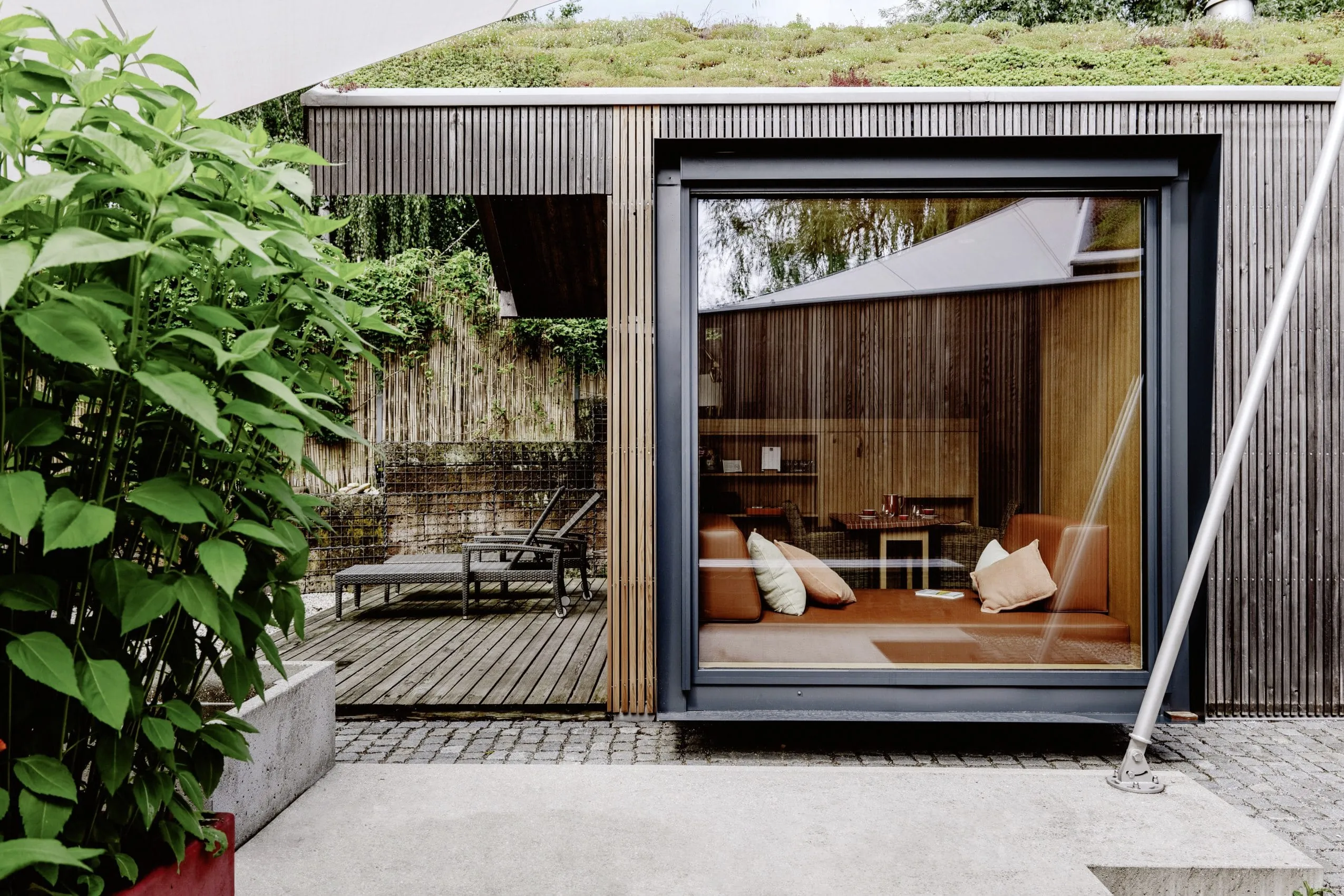 Blick auf und in das Hanghaus aus Holz auf dem Hofgut Hafnerleiten mit gem&uuml;tlichem Sofa direkt am gro&szlig;en Panoramafenster und Liegest&uuml;hlen auf der Terrasse