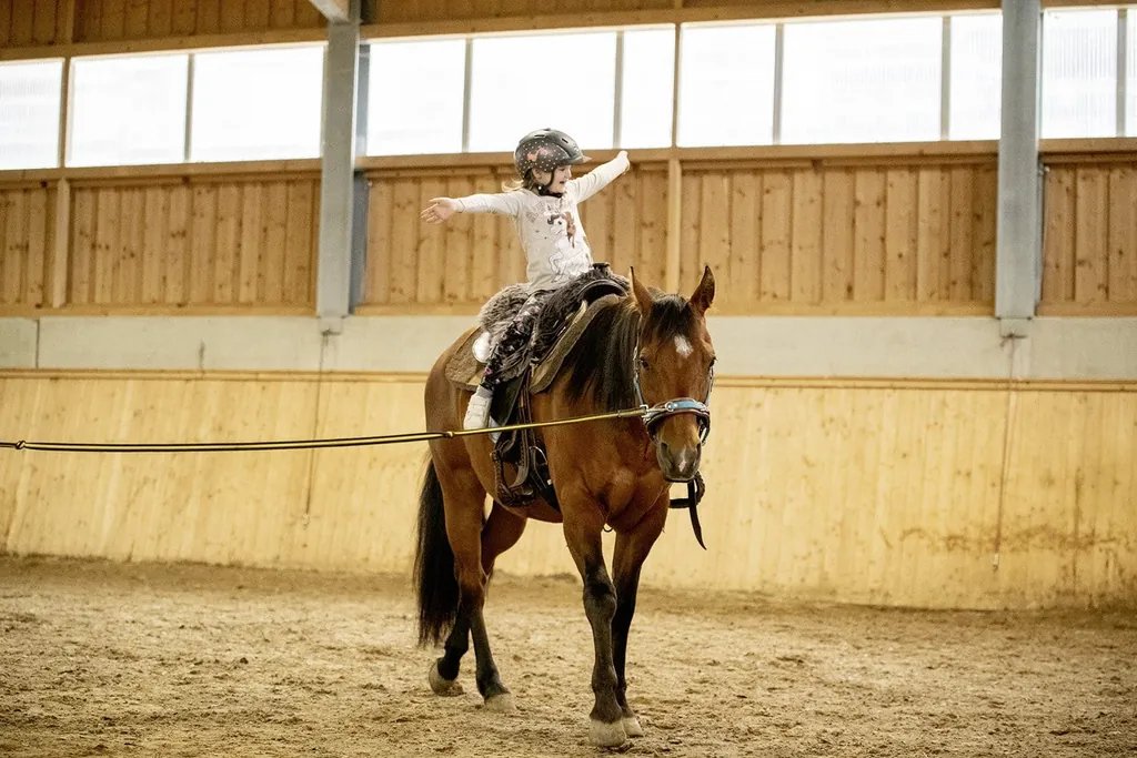 M&auml;dchen sitzt auf einem braunen Pferd in der Reithalle des Hotel & Resort Landhaus zur Ohe und streckt die Arme aus