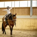 Mädchen sitzt auf einem braunen Pferd in der Reithalle des Hotel & Resort Landhaus zur Ohe und streckt die Arme aus