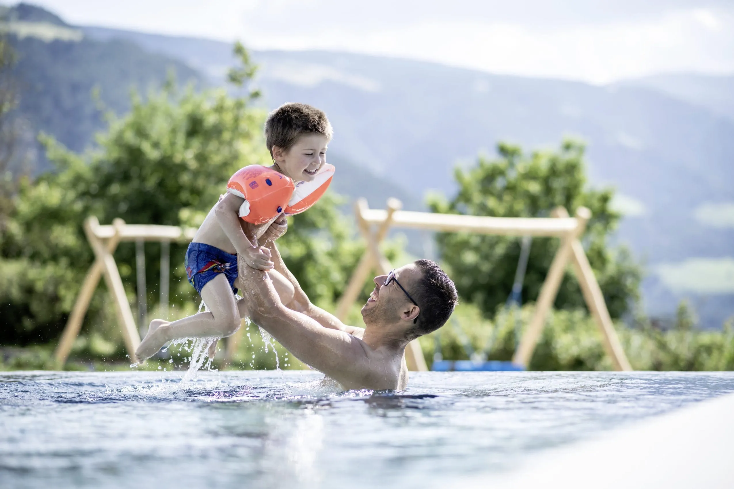 Mann mit Brille wirft seinen Sohn mit Schwimmflügeln im Pool des Das Mühlwald in die Luft