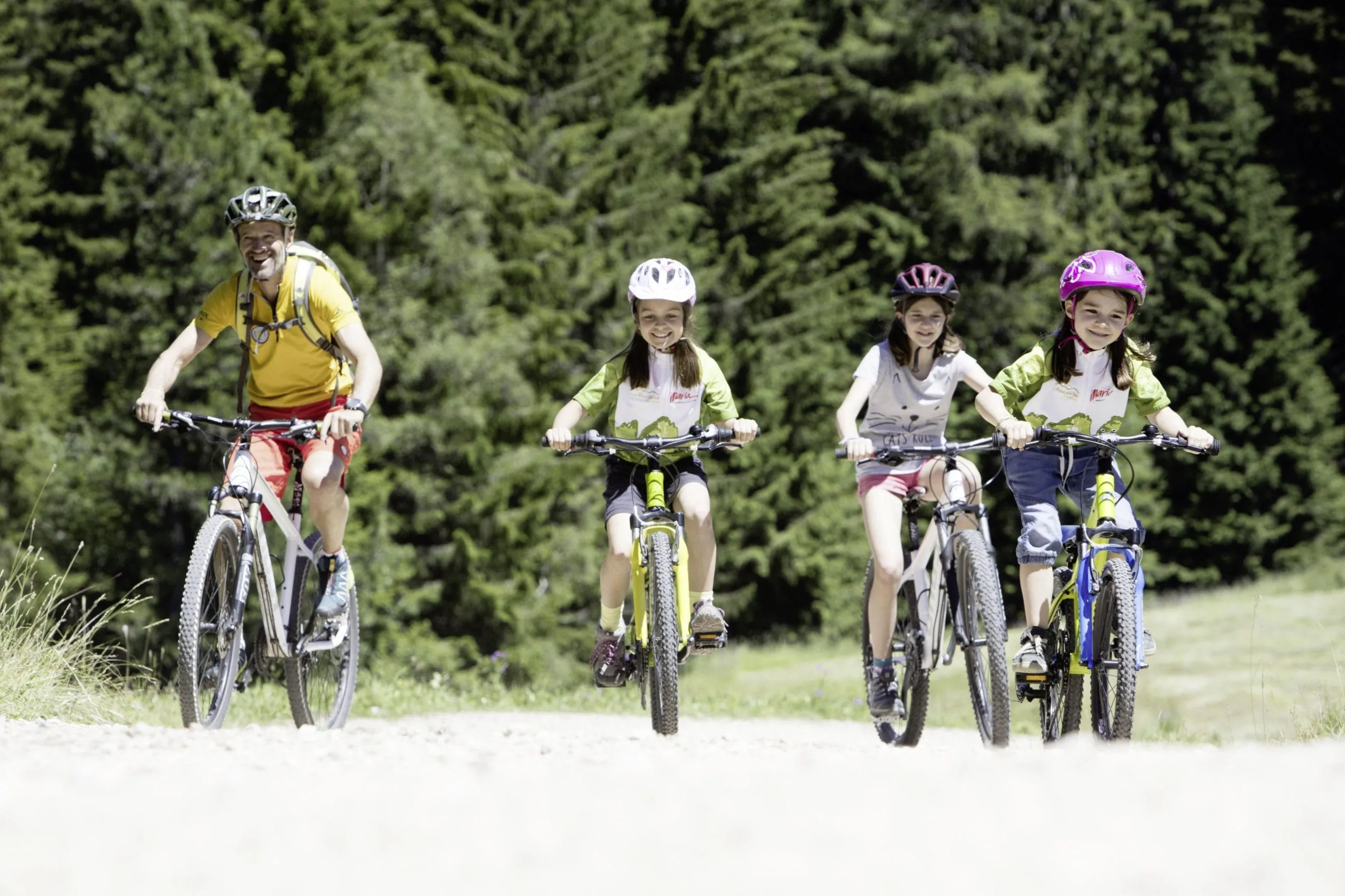 Mann mit gelbem T-Shirt fährt im Adventure Family Hotel Maria mit drei Mädchen mit Helm Fahrrad