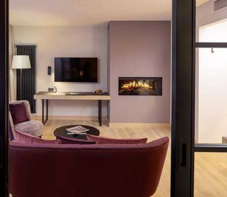 Blick vom Schlafzimmer einer Suite des Hotel Engel Obertal ins Wohnzimmer mit roten Sitzmöbeln, Flachbildfernseher und Kaminfeuer