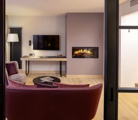 Blick vom Schlafzimmer einer Suite des Hotel Engel Obertal ins Wohnzimmer mit roten Sitzmöbeln, Flachbildfernseher und Kaminfeuer