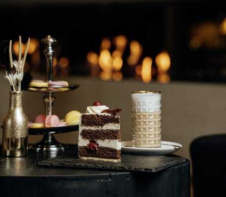großes Stück Schwarzwälder Kirschtorte auf einer Schieferplatte, Kaffeegetränk und Macarons im Hintergrund