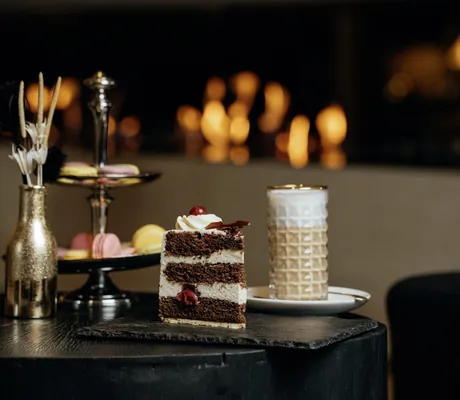 großes Stück Schwarzwälder Kirschtorte auf einer Schieferplatte, Kaffeegetränk und Macarons im Hintergrund