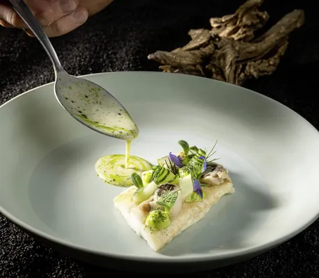 Koch gießt helle Soße mit einem Löffel auf einen mit einer Seezunge schön angerichteten tiefen Teller