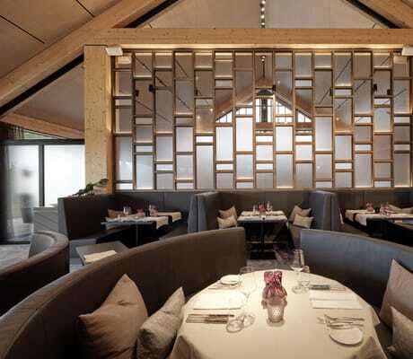Blick in das Restaurant "Esszimmer" des Öschberghofs mit modernen, dunkelgrauen Sitzbänken, gedeckten Tischen und großer Trennwand aus Holz mit Glasdetails