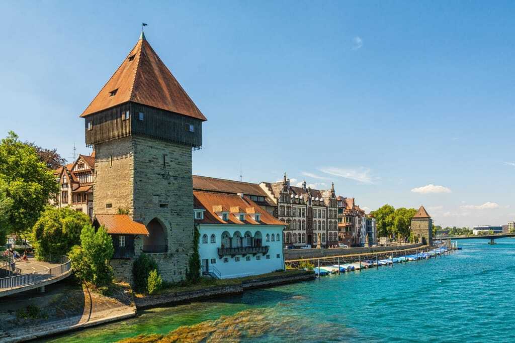 Der Rheintorturm in Konstanz am Bodensee, direkt an der Uferpromenade. Daneben historische Fachwerkhäuser und der glitzernd-blaue Bodensee. 