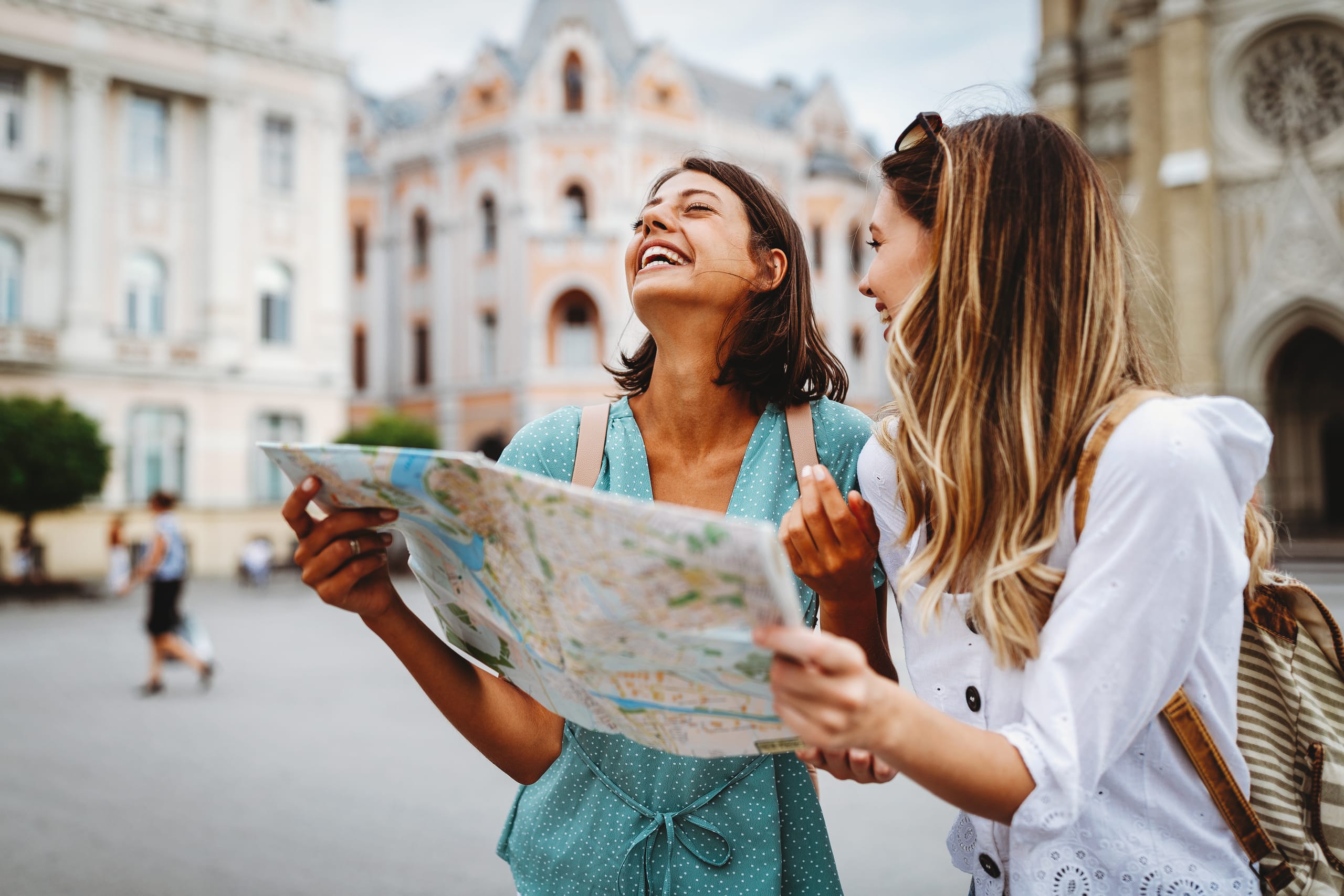 Zwei junge Frauen machen einen Städtetrip in Süddeutschland und stehen lachend mit einer Karte in der Hand auf einem Stadt-Marktplatz.