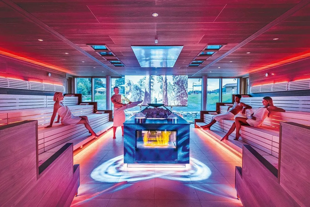 Gro&szlig;e Sauna mit stimmungsvoller Beleuchtung und einem zentralen Saunaofen in der Mitte. Mehrere G&auml;ste entspannen auf den breiten Sitzb&auml;nken.