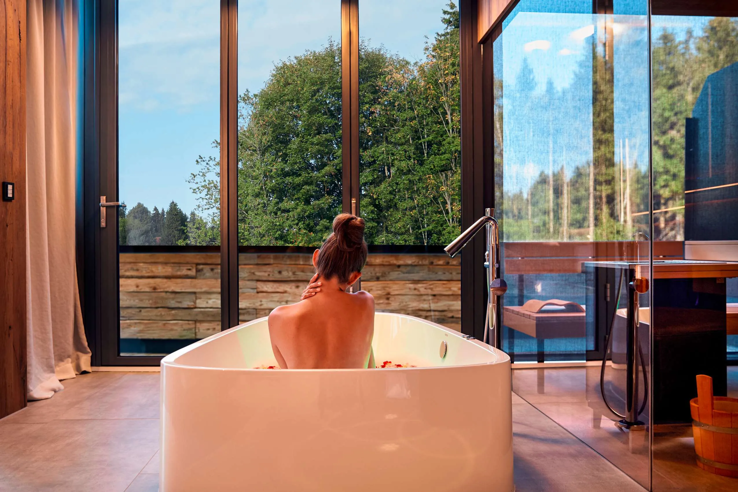 Frau in einer freistehenden Badewanne vor gro&szlig;en, bodentiefen Fenstern.