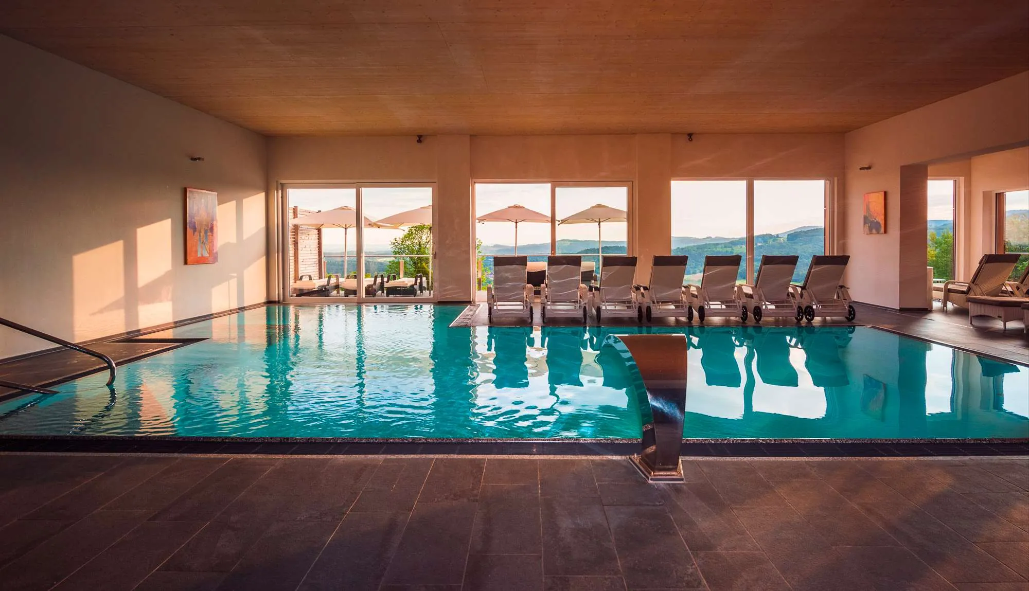 Hallenbad mit Liegen und Panoramafensterfront mit Ausblick auf die h&uuml;gelige Landschaft