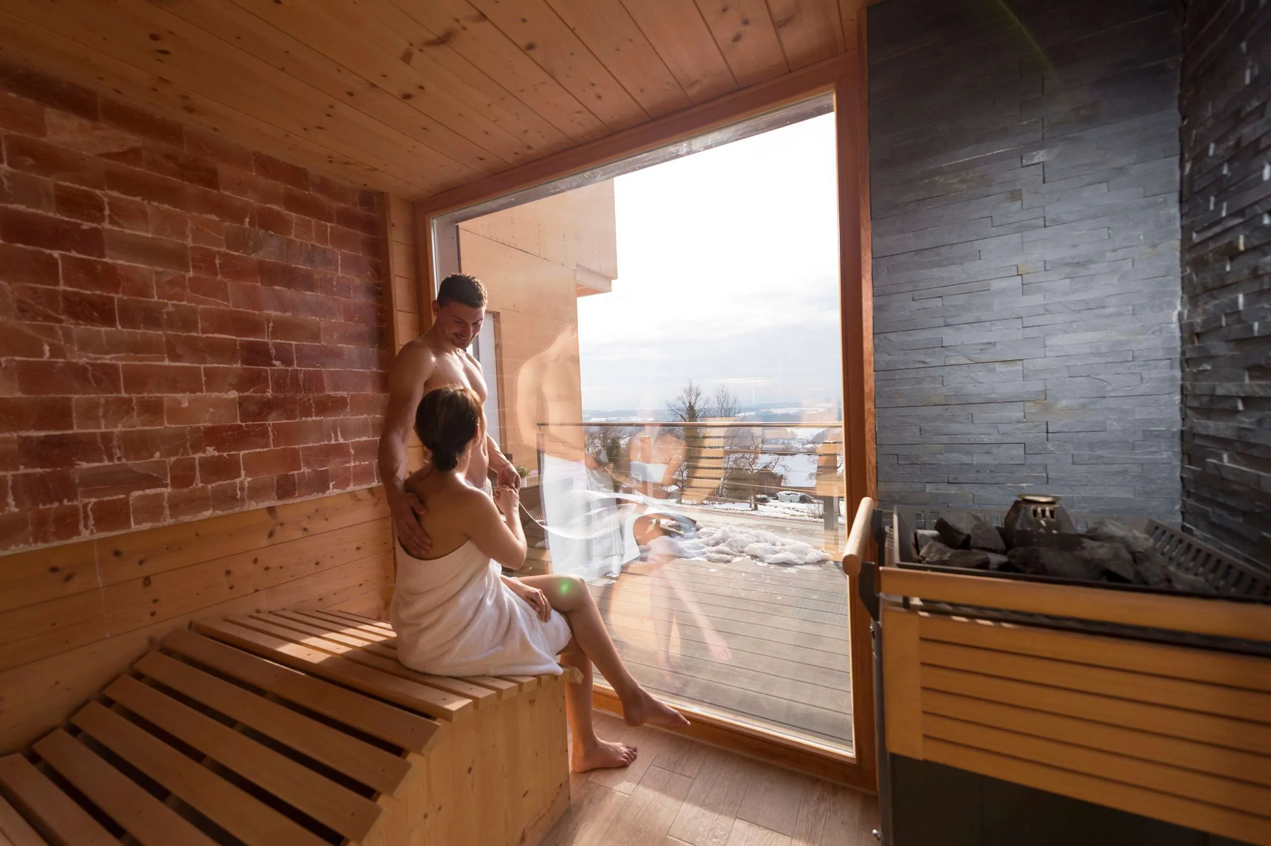 Sauna mit dunklen Natursteinflie&szlig;en und rotem Backstein an den W&auml;nden, sowie gro&szlig;em Fenster nach drau&szlig;en auf den Balkon.