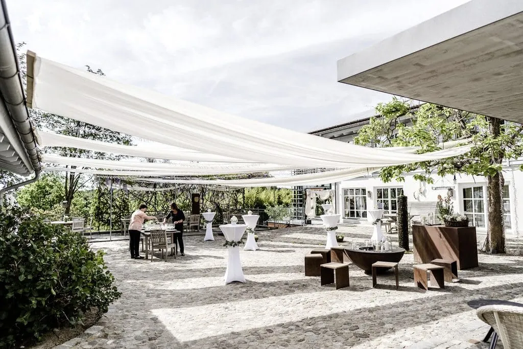 Gepflasterter Innenhof umgeben von viel Gr&uuml;n, &uuml;ber den wei&szlig;e Stoffbahnen als Sonnensegel gespannt wurden und elegante Stehtische darunter stehen.