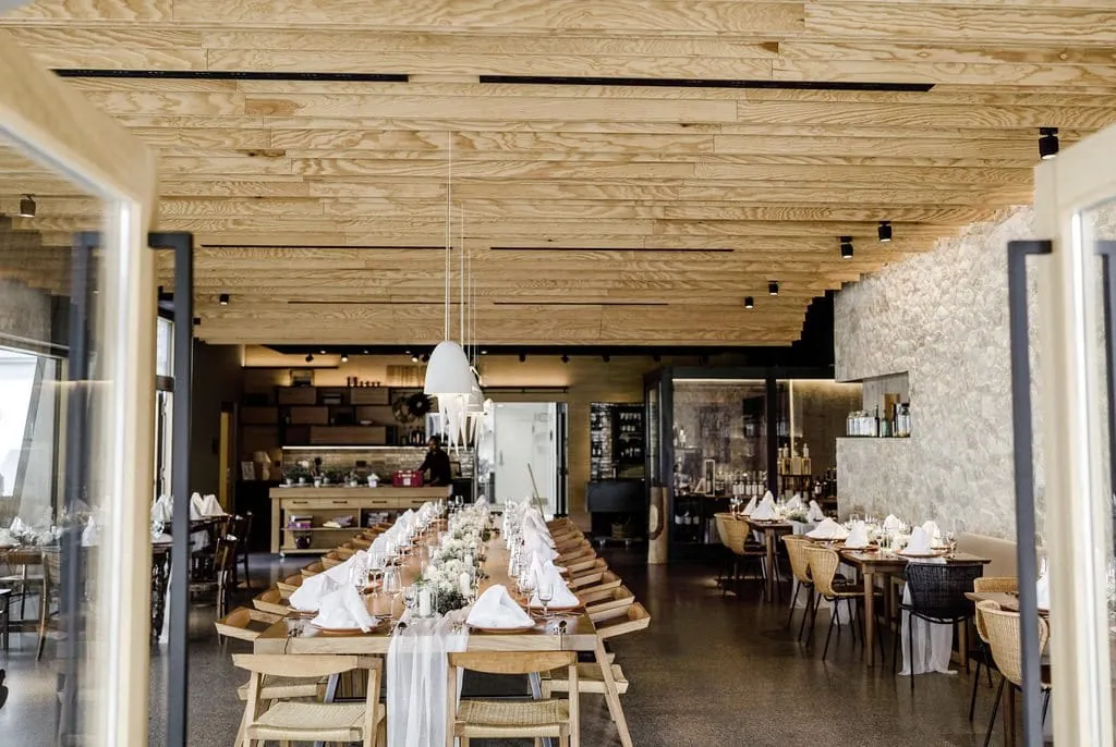 Glashaus des Hofgut Hafnerleiten mit Holztischen und St&uuml;hlen in warmen T&ouml;nen, dekoriert mit wei&szlig;en Stoffservietten und Blumengestecken.