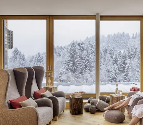 Paar in weißen Bademänteln sitz in Ohrensesseln vor dem Panoramafenster mit Blick auf verschneite Landschaft