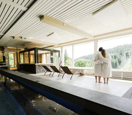 Pärchen in weißen Bademänteln steht umarmt am Beckenrand des Hallenbades im Waldhotel am Notschreipass