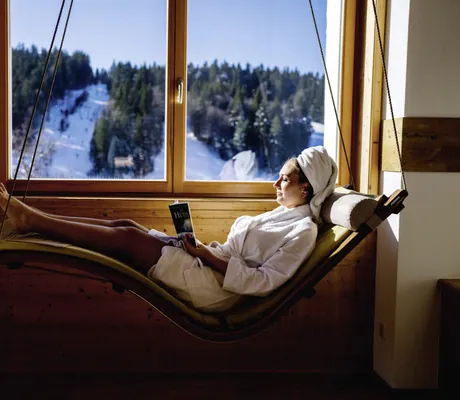 Frau im Bademantel und Frotteeturban liegt auf der Hängeliege am Fenster des Waldhotel am Notschreipass