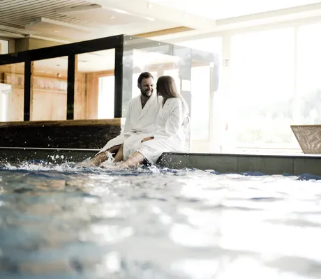 Paar in weißen Bademänteln sitzt am Beckenrand des Hallenbades im Waldhotel am Notschreipass